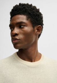 Beiger Pullover aus strukturiertem Stoff, mit runder Halslinie und geripptem Saum, getragen von einem Modell mit kurzen, lockigen Haaren.