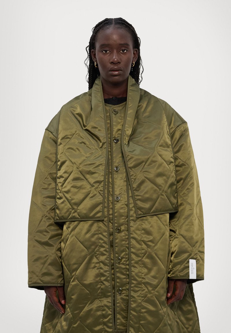 Olivgrüne gestepte Pufferjacke mit abnehmbarem Kragen, ausgestattet mit Druckknöpfen, einem lässigen Schnitt und subtilem Texturmuster im gesamten Kleidungsstück.