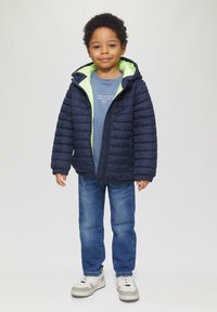 Marineblauwe pufferjack met een felgroene binnenkant, rits aan de voorkant, geribde manchetten, gedragen over een blauw T-shirt en gecombineerd met blauwe spijkerbroek en witte sneakers.