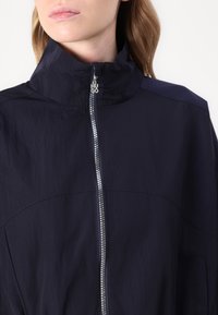 Giacca zip-up blu navy con colletto alto, dotata di una cerniera argentata e logo in rilievo. Il tessuto sembra leggero e ha una texture liscia.