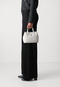 Geantă albă cu un model, având o formă rotundă, decorată cu accente negre și două mânere scurte. Include un detaliu logo triunghiular pe față.