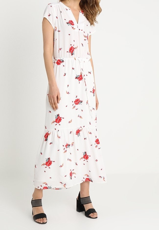 Femme portant une robe midi blanche à fleurs avec des roses rouges, manches courtes, col en V et ceinture blanche à la taille, associée à des sandales à talons noires.