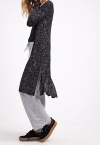 Pitkä musta kardigan, jossa on tekstuuri kuvio, avoin etuosa, sivuhalkiot; yhdistetty harmaisiin teksturoituihin housuihin ja mustiin lenkkareihin, joissa on kumipohjat.