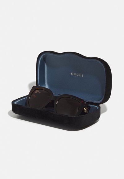 Černé sluneční brýle Gucci v měkkém černém a modrém pouzdře s zlatým logem. Brýle mají výrazný čtvercový tvar a tmavé čočky.