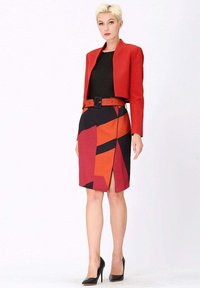 Veste rouge courte sur un haut noir, associée à une jupe crayon à motif géométrique multicolore avec une fente à l'avant, une ceinture noire et des chaussures à talons.