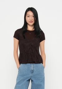 Een bruine kanten peplumtop met korte mouwen, een v-hals en knoopsluiting, gedragen over een donkere bordeauxkleurige, aansluitende shirt. Gecombineerd met lichtblauwe wijde jeans.