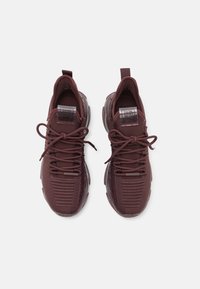 Paire de baskets tricotées marron foncé avec des bouts ronds, des languettes au niveau des talons et des semelles épaisses texturées, vues de dessus sur un fond blanc.