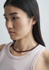 Halskæde lavet af mørkerøde perler med uregelmæssig form, båret over en lyserød ærmeløs top, hvilende på halsudskæringen. Blank tekstur.