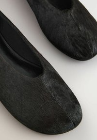 Paire de chaussures plates noires à enfiler avec une tige lisse, texturée comme de la fourrure, et des bouts arrondis sur fond clair.
