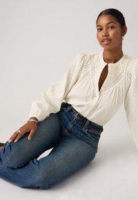 Γυναίκα με σκούρα μαλλιά σε χαμηλό σινιόν φοράει λευκό μπλουζάκι με λεπτομέρειες από δαντέλα στα μανίκια και ψηλόμεσα μπλε τζιν, καθισμένη στο πάτωμα.