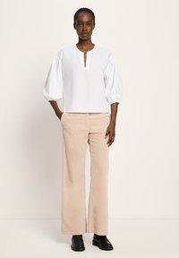 Frau in einer weißen Bluse mit gepufften Ärmeln, beigen weiten Hosen und schwarzen Slippern, die mit Händen in den Taschen vor einem einfarbigen Hintergrund steht.