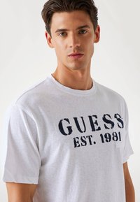 Weißes Baumwoll-T-Shirt mit Rundhalsausschnitt und kurzen Ärmeln, mit schwarzem "GUESS EST. 1981" Grafikdruck über der Brust.