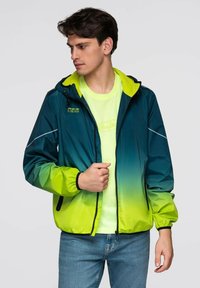 Mladý muž v bundě s zelenožlutým gradientem, pod kterou má na sobě neonově žluté tričko a modré džíny, se dívá doleva na jednobarevném pozadí.