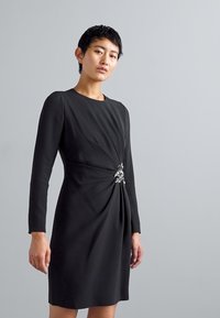 Abito lungo nero con maniche lunghe e scollo rotondo, caratterizzato da tessuto arricciato in vita e un accento decorativo di strass. Texture liscia.