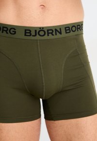 Olivengrønne boxershorts med en sort elastisk talje, der har "BJÖRN BORG" trykt på. Glat tekstur, lår-længde, dobbeltsøm.