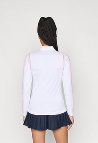 Valkoinen pitkähihaisella urheilupaita, jossa on pinkkejä yksityiskohtia, pyöreä kaulus ja verkkomaiset sivupaneelit, yhdistettynä tummansinihen pleatattuun hameeseen.