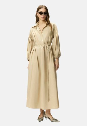 Donna che indossa un abito beige a maniche lunghe con cintura, spalle testurizzate, occhiali da sole neri a forma di occhio di gatto, orecchini a cerchio dorati e scarpe a punta in pelle di serpente.