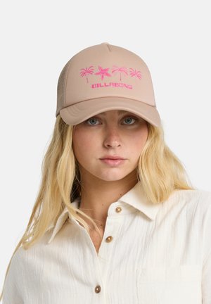 Młoda kobieta o długich blond włosach nosi beżową czapkę Billabong z różowym wzorem palm i rozgwiazd oraz białą koszulę na guziki.