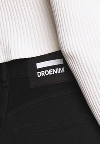 Jeans de mezclilla negra con un parche del logo de DRDENIM visible. La parte superior es de una tela ligera y con textura acanalada. La vista en primer plano resalta la textura y las costuras.