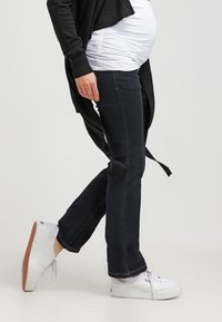 Graviditetsjeans i mörk tvättad denim, rak ben-design med mid-rise midja, kombinerat med en vit topp och vita sneakers.