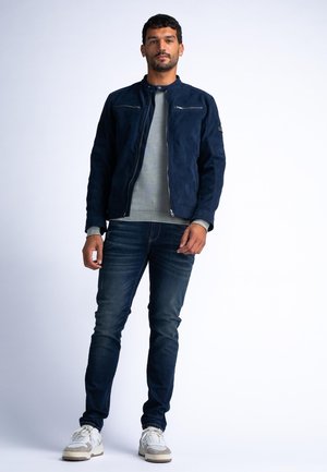 Navy suède jack met een ronde kraag, voorzijde met ritssluiting en twee ritssluitingen op de borst, gecombineerd met een grijze sweater, donkere jeans en witte sneakers.