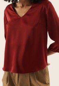 Blouse rouge à manches courtes avec un col en V, tissu doux et légèrement brillant, avec une petite poche sur le côté gauche. Associée à un pantalon beige.