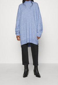 Ljusblå oversized tröja med hög krage, långa ärmar och slits i nederkant, tillsammans med svarta tajta jeans och ankellånga stövlar.