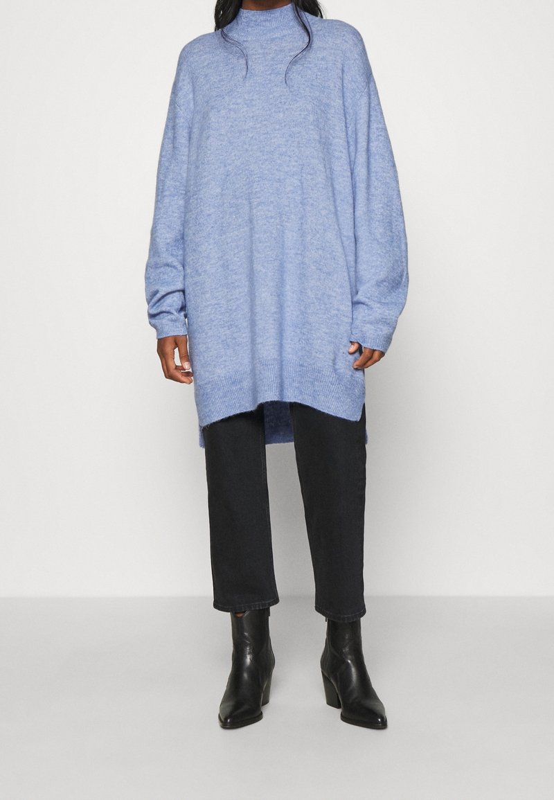 Ljusblå oversized tröja med hög krage, långa ärmar och slits i nederkant, tillsammans med svarta tajta jeans och ankellånga stövlar.