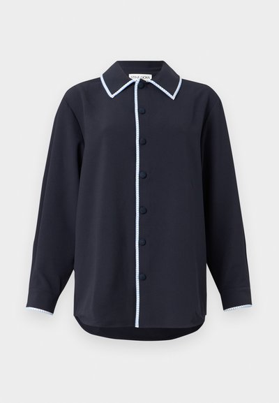 Tumši zila pogājama krekla ar garām piedurknēm, ar kontrastējošu gaiši zilu apmali apkārt apkaklei un pogu lentei, kā arī ar izliektu apakšmalu.