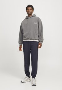 Grå hættetrøje med frontlomme, båret over en hvid skjorte. Sammen med navy sweatpants og sølv atletiske sko. Simpelt design, afslappet pasform.