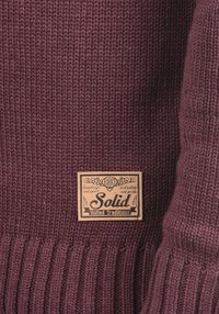 Bordeauxroter Strickpullover mit gerippter Struktur und einem rechteckigen Lederetikett mit der Aufschrift „Solid“ und dekorativem Text, angebracht unten links.