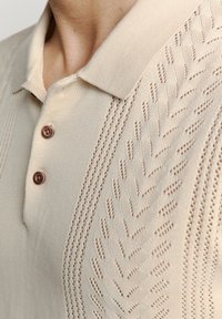 Beige polo t-shirt met een gebreid patroon, voorzien van een geribbelde kraag en twee bruine knopen op de sluiting. Zachte textuur en ademend ontwerp.