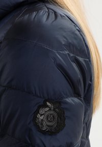 Manche de veste matelassée bleu foncé avec un patch circulaire noir brodé des lettres IKKKS, portée par une personne aux longs cheveux blonds.