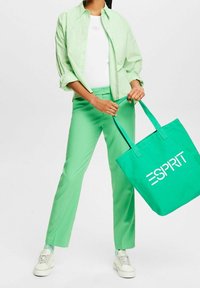 Light green button-up shirt, white t-shirt, and green pants. Holding a turquoise tote bag with "ESPRIT" logo in bold white letters.
