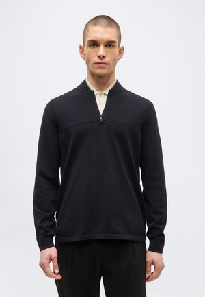 Młody mężczyzna z krótkimi włosami, ubrany w czarny sweter zapinany do ćwiartki na jasną koszulę z kołnierzykiem, stoi na tle jednolitego białego tła.