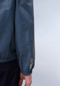 Donkerblauwe lichtgewicht jas met elastische manchetten, voorzien van een logo patch met het label "CORDURA" op de mouw, gemaakt van duurzaam stof.