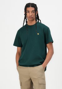 Green cotton t-shirt with a crew neckline, short sleeves, and a small yellow logo on the chest. Worn with khaki cargo pants.