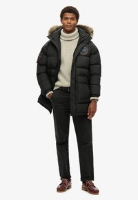 Chaqueta de abrigo negra con capucha forrada de piel, tela texturizada y parche en el pecho. Lleva puesta sobre un suéter de cuello alto gris claro y pantalones negros.