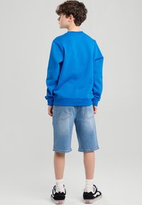 Sininen pitkähihaisen huppari, joka on yhdistetty vaaleansinisiin polvenpituisiin farkkushortseihin. Asusteina valkoiset sukat ja mustat lenkkarit.