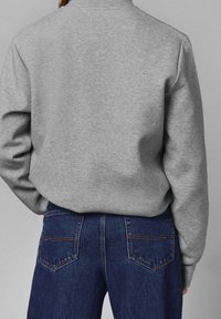 Szara bluza z okrągłym dekoltem, noszona na ciemnoniebieskich dżinsach z tylnymi kieszeniami i widocznym detalem szwów.