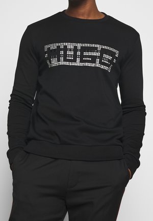Person wearing a black long-sleeve shirt with "HUGO" in silver block letters made of square tiles on the chest.