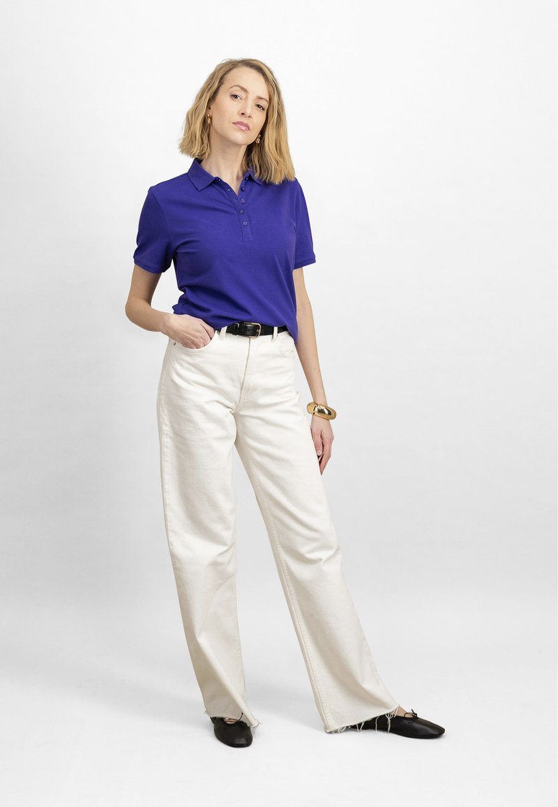 Sieviete violetā polo krekliņā, baltās augsta vidukļa plaši piegrieztas biksēs, melnā jostā, zelta aprocē un melnās kurpēs, stāvot pret baltu fonu.