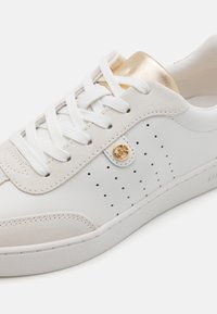 Witte sneaker van leer en suède, met een geperforeerd ontwerp, gouden accenten op de hak en witte veters. Dikke rubberen zool.