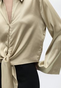 Μπεζ σατέν μπλούζα με βαθύ V λαιμόκοψη και λεπτομέρεια κόμπου μπροστά, φορεμένη με μαύρο παντελόνι με υψηλή μέση.