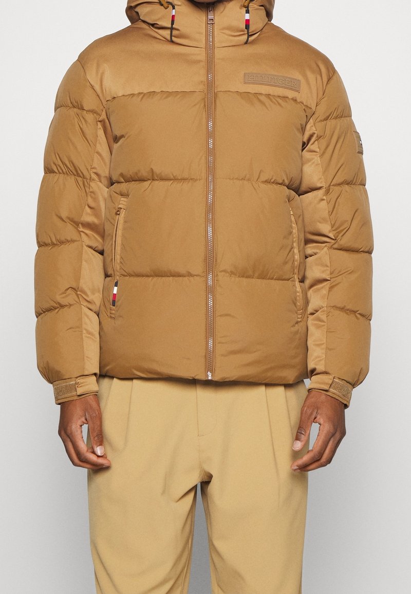 Brown puffer jacket with a hood, featuring a front zipper, elastic cuffs, and logo patch. Smooth texture with quilted design and side pockets.