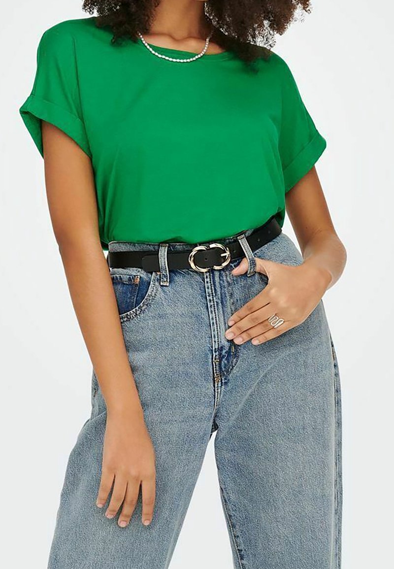 Femme portant un haut vert à manches courtes rentré dans un jean bleu taille haute avec une ceinture noire et une boucle argentée.