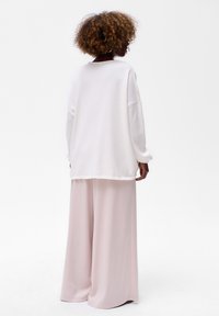 Persoon met krullend haar die een oversized witte trui en brede lichtroze broeken draagt, met de rug naar de camera gekeerd tegen een witte achtergrond.