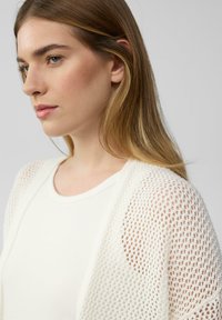 Witte opengebreide cardigan over een crèmekleurige tanktop, met een textuurpatroon en een losse pasvorm, voorzien van een ronde halslijn en subtiele accenten.
