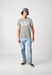 Tricou gri din bumbac cu logo mare alb "GAP" și accente roșii, asortat cu blugi denimi de un bleu deschis și adidași negri cu talpă albă.