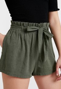 Vihreät pellavashortsit rentolla istuvuudella, joissa on joustava vyötärönauha, sivutaskut ja solmittava vyötärönauha. Sileä pinta, polvipituus.